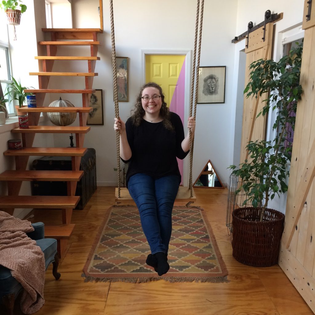Cara sits on a swing in front of a yellow and pink door in the middle of an apartment decorated with plants. She is smiling. 